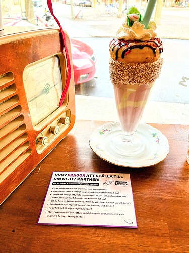 En gammaldags radio står bredvid en imponerande milkshake med dekorationer och bredvid ligger en <tag>note</tag> med frågor att ställa på dejt eller till en partner. (Captioned by AI)