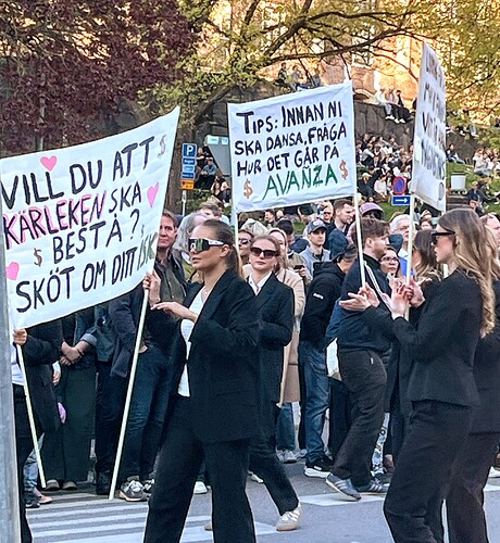 En grupp människor deltar i en protest eller demonstration, där flera kvinnor klädda i svart kostym bär skyltar med svenska budskap. (Captioned by AI)